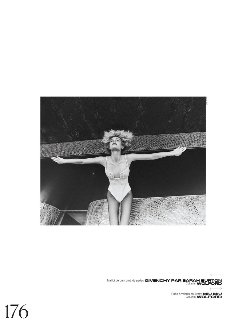 Black and white fashion shot of a blond woman wearing a bodysuit with arms outstretched, against a dark mosaic-tiled surface.