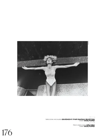 Black and white fashion shot of a blond woman wearing a bodysuit with arms outstretched, against a dark mosaic-tiled surface.