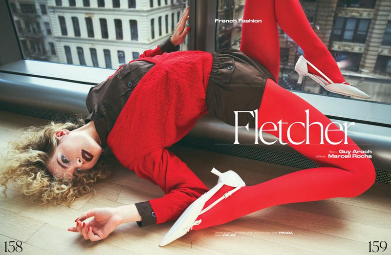 Fashion editorial shot of a model in red tights and white heels, lying on a wood floor in front of a city view window.