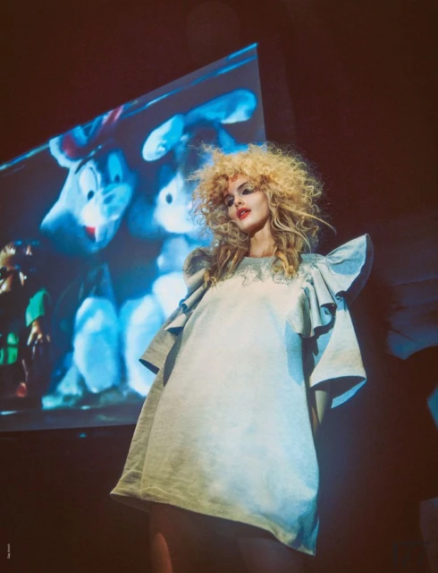 A woman with blonde curly hair stands in a light blue dress with a projection of cartoon animals displayed behind her.