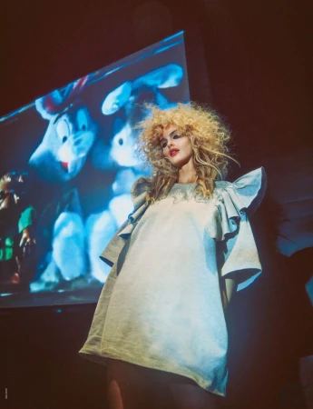 A woman with blonde curly hair stands in a light blue dress with a projection of cartoon animals displayed behind her.