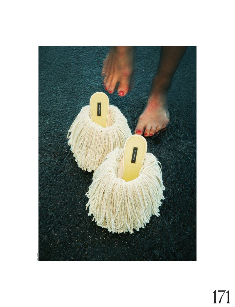 Novelty shoes with yarn fringe stand on a textured surface, accompanied by bare feet with red nail polish in a studio setting.