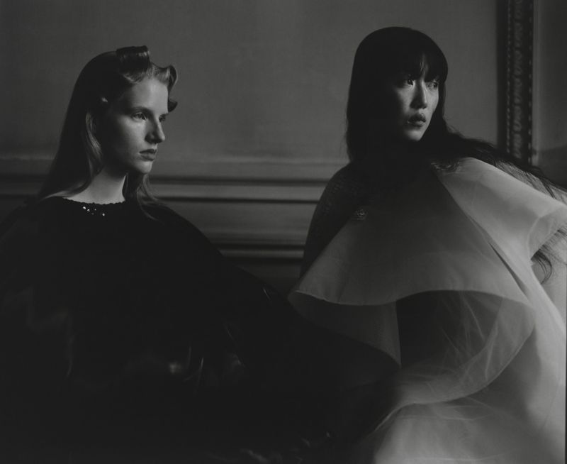 Monochrome studio portrait of two women with dark hair in elegant draped gowns against a neutral background with classic trim.