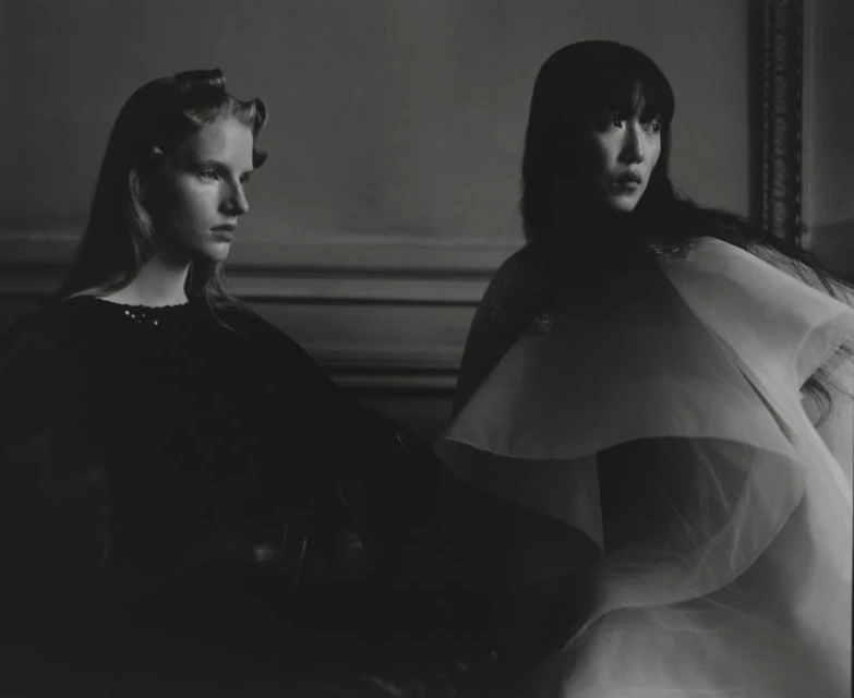 Monochrome studio portrait of two women with dark hair in elegant draped gowns against a neutral background with classic trim.
