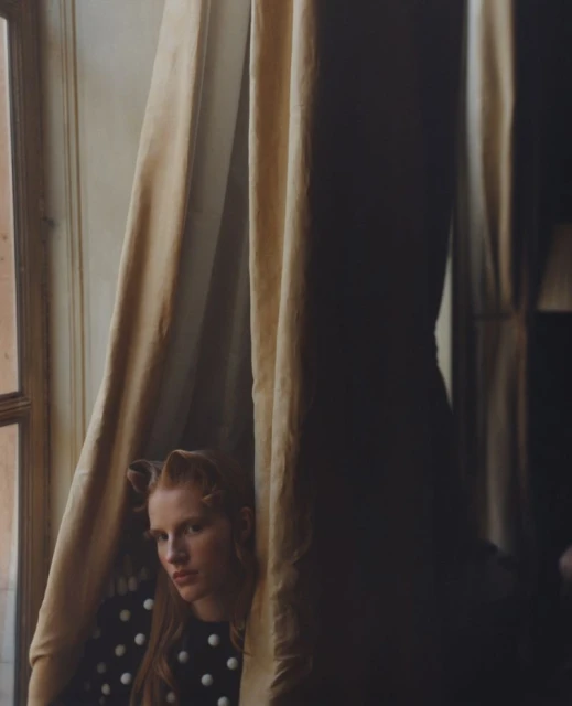 Woman with red hair sits by a window with cream-colored curtains, wearing a black top with white polka dots in an indoor setting.