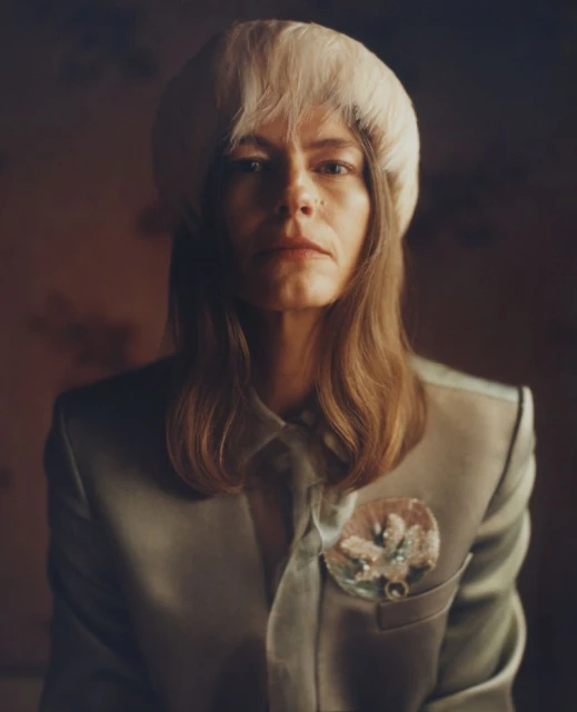 Close-up portrait of a woman with long blonde hair and a cream-colored hat, wearing a light green suit with a floral pin.