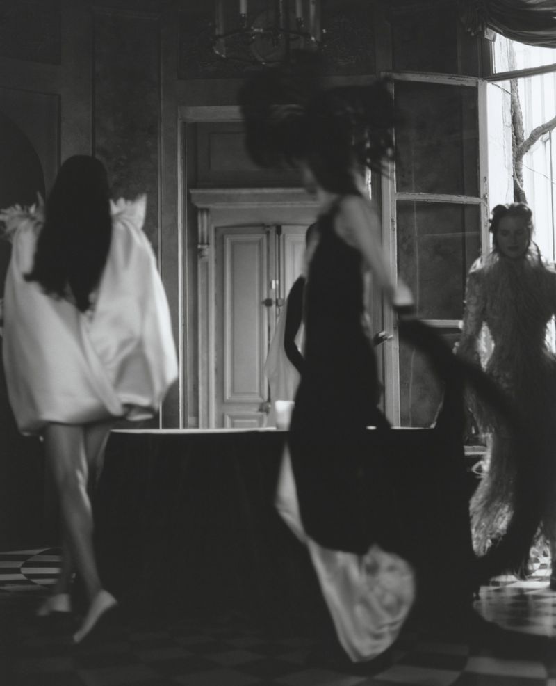 Three women in a black and white fashion editorial, wearing elegant dresses, dancing in a grand interior with a chandelier and large windows.