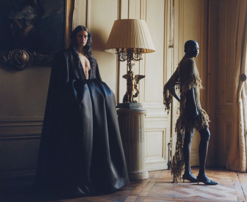 Two fashion models in elegant gowns pose indoors near a lamp. One model wears a dark gown, while the other is in a light-colored gown.