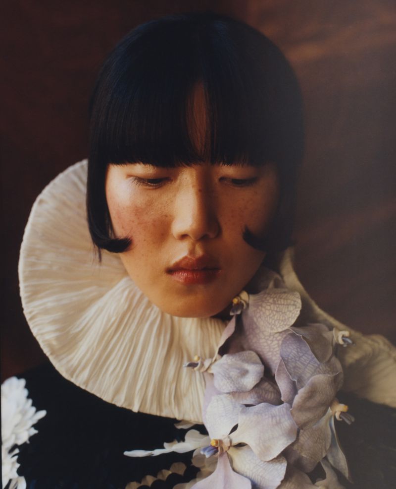 Portrait of an Asian woman with a dark blunt bob and a white ruffled collar, accentuated by a flower, set against a blurred brown background.