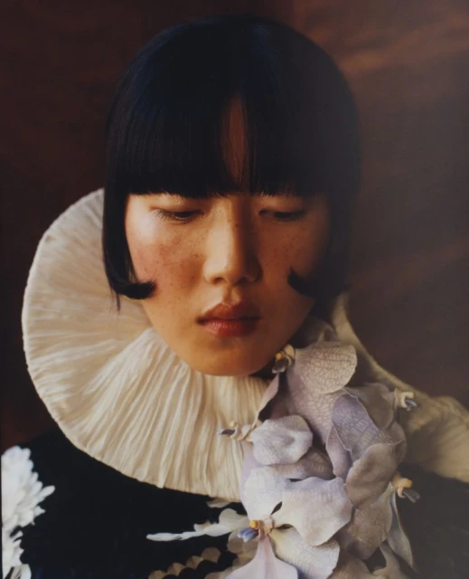 Portrait of an Asian woman with a dark blunt bob and a white ruffled collar, accentuated by a flower, set against a blurred brown background.