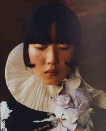 Portrait of an Asian woman with a dark blunt bob and a white ruffled collar, accentuated by a flower, set against a blurred brown background.
