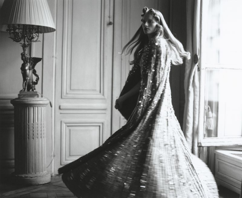 Black and white image of a woman in a shimmering gown twirling in an ornate room with a lamp and window, fashion photography.