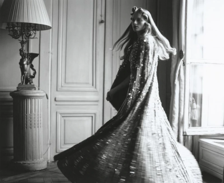 Black and white image of a woman in a shimmering gown twirling in an ornate room with a lamp and window, fashion photography.