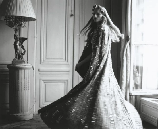 Black and white image of a woman in a shimmering gown twirling in an ornate room with a lamp and window, fashion photography.