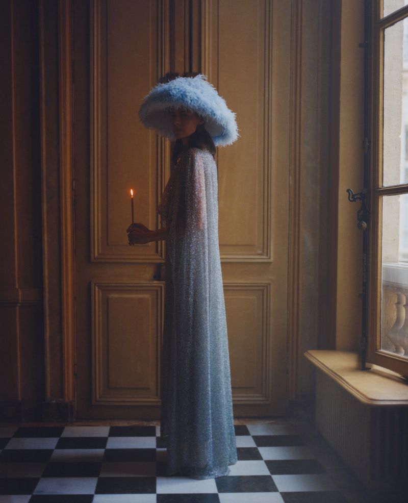 Elegant woman in sparkly gown and large feathered hat holding a candle, standing in a room with checkered floor and paneled walls.