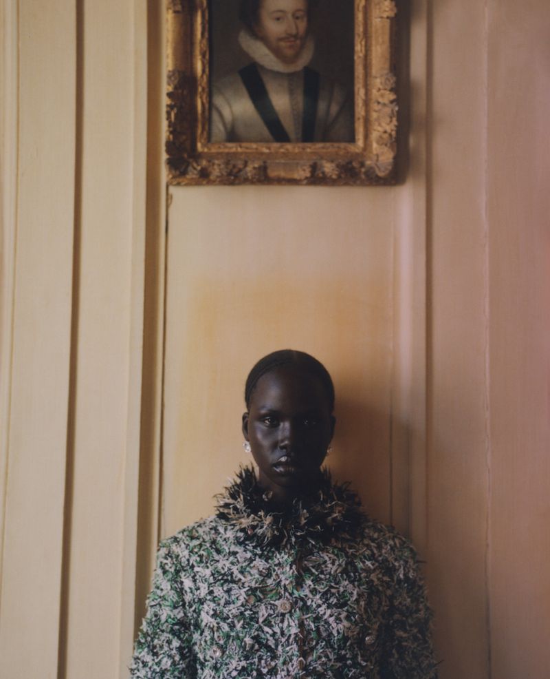 Portrait of a dark-skinned woman in a green and white tweed jacket with a portrait in gold frame hanging above on a neutral wall.
