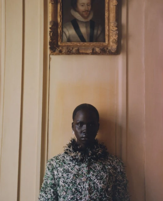 Portrait of a dark-skinned woman in a green and white tweed jacket with a portrait in gold frame hanging above on a neutral wall.