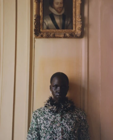 Portrait of a dark-skinned woman in a green and white tweed jacket with a portrait in gold frame hanging above on a neutral wall.