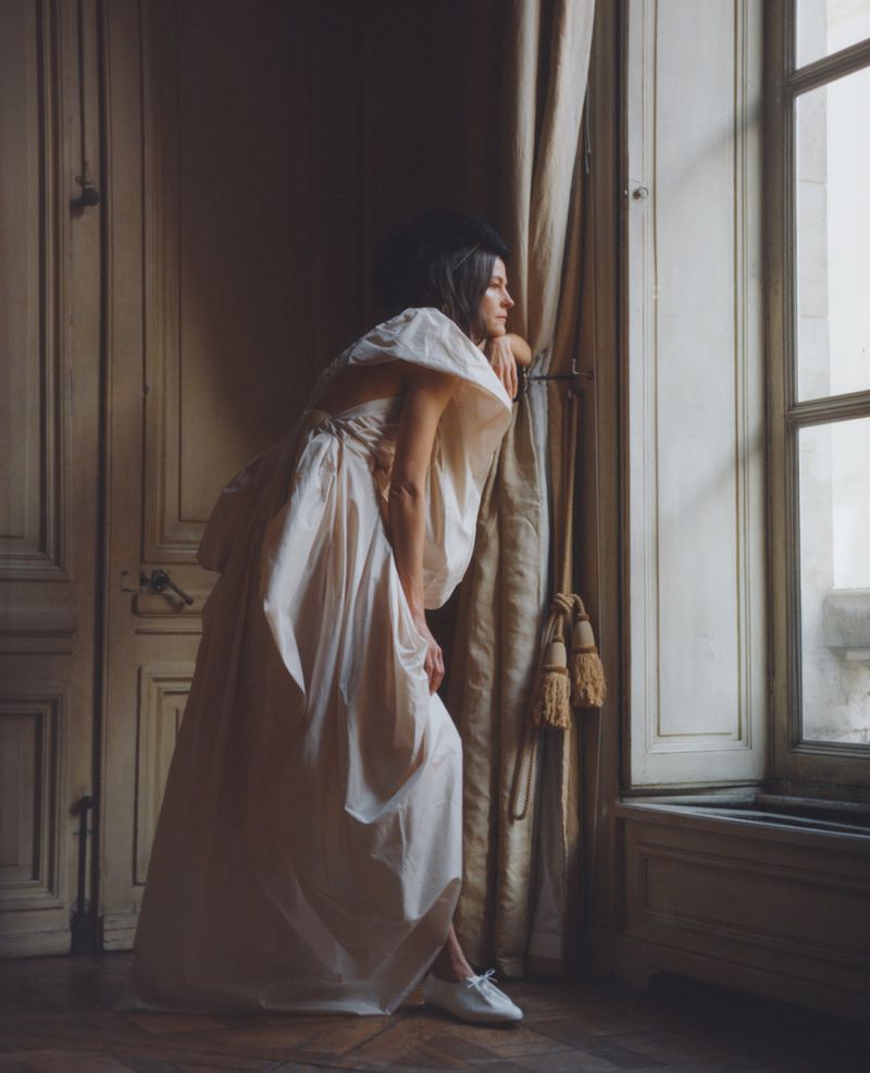 A woman in an off-white gown looking out of a window in a vintage room with wood paneled doors and walls and draped curtains.