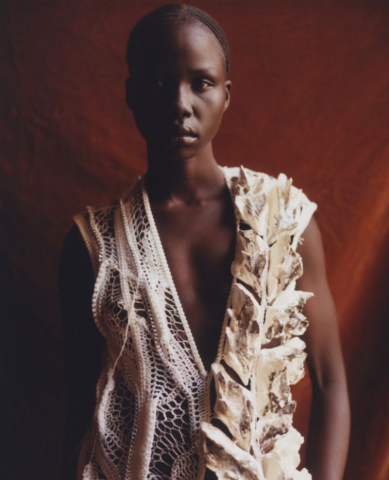 Portrait of a dark-skinned woman wearing a decorative vest against a textured reddish-brown backdrop in soft, diffused lighting.