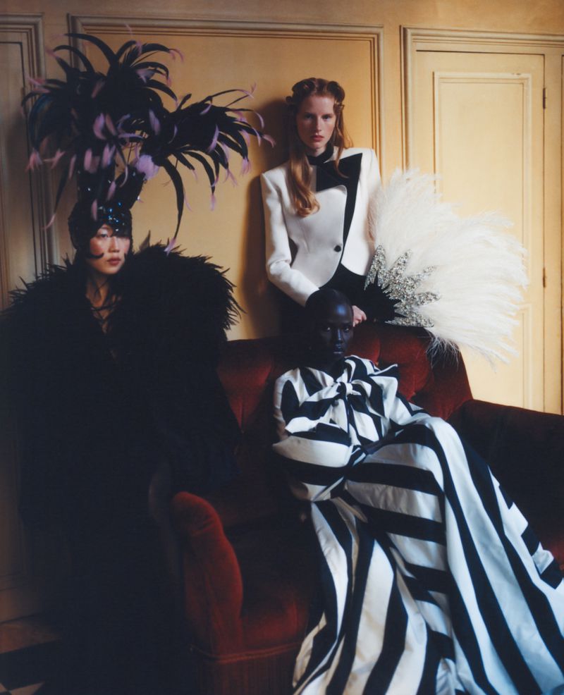 Four fashion models pose together in an indoor setting, wearing elegant couture with dramatic feathered headpieces and apparel with stripes and bows.