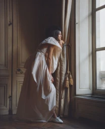 Model wearing a voluminous white gown standing by a large window in a classic interior by Antonino Cafiero for Vogue Polska.