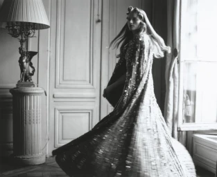 Model in a sweeping sequined gown turning gracefully inside a room with a decorative floor lamp and panelled doors.