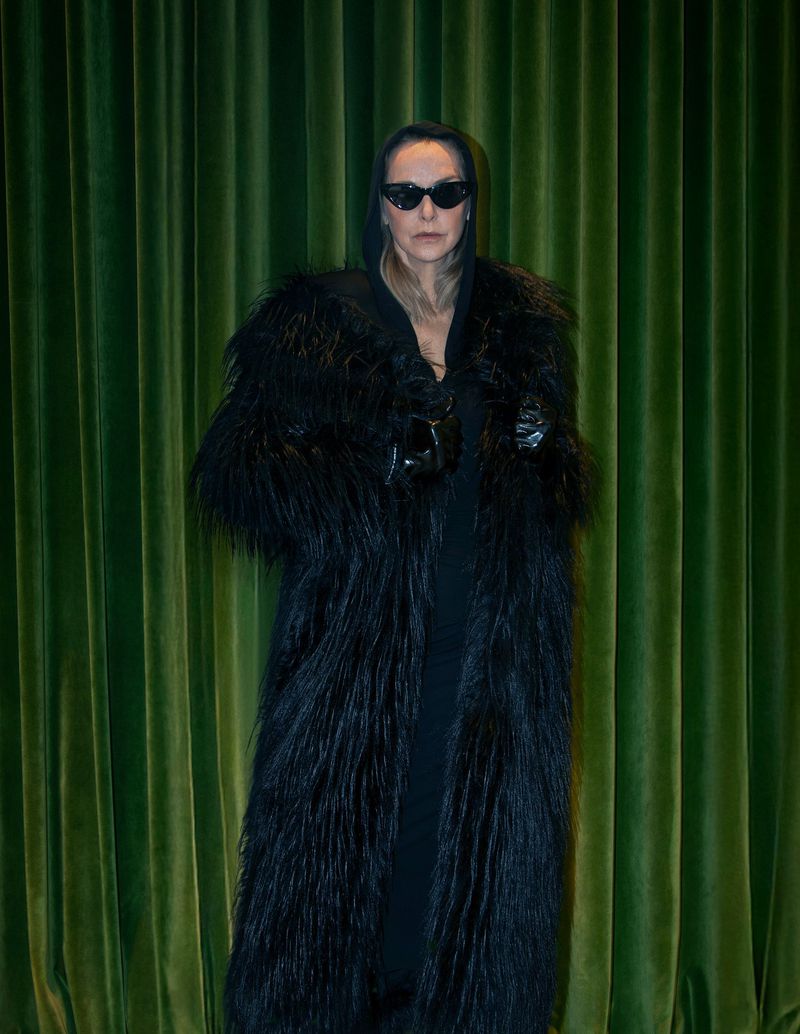 Adult female wearing a long black fur coat and sunglasses stands against a green curtain backdrop in a studio setting.