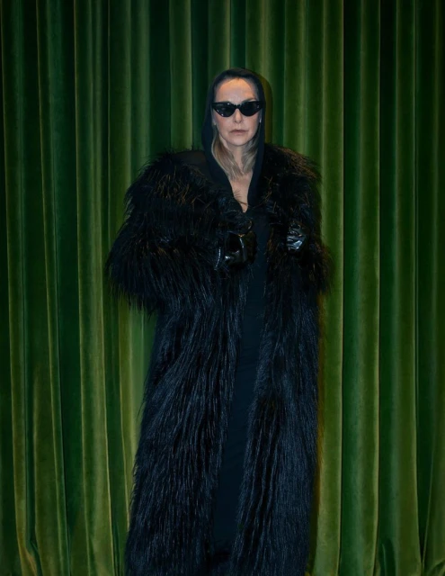 Adult female wearing a long black fur coat and sunglasses stands against a green curtain backdrop in a studio setting.