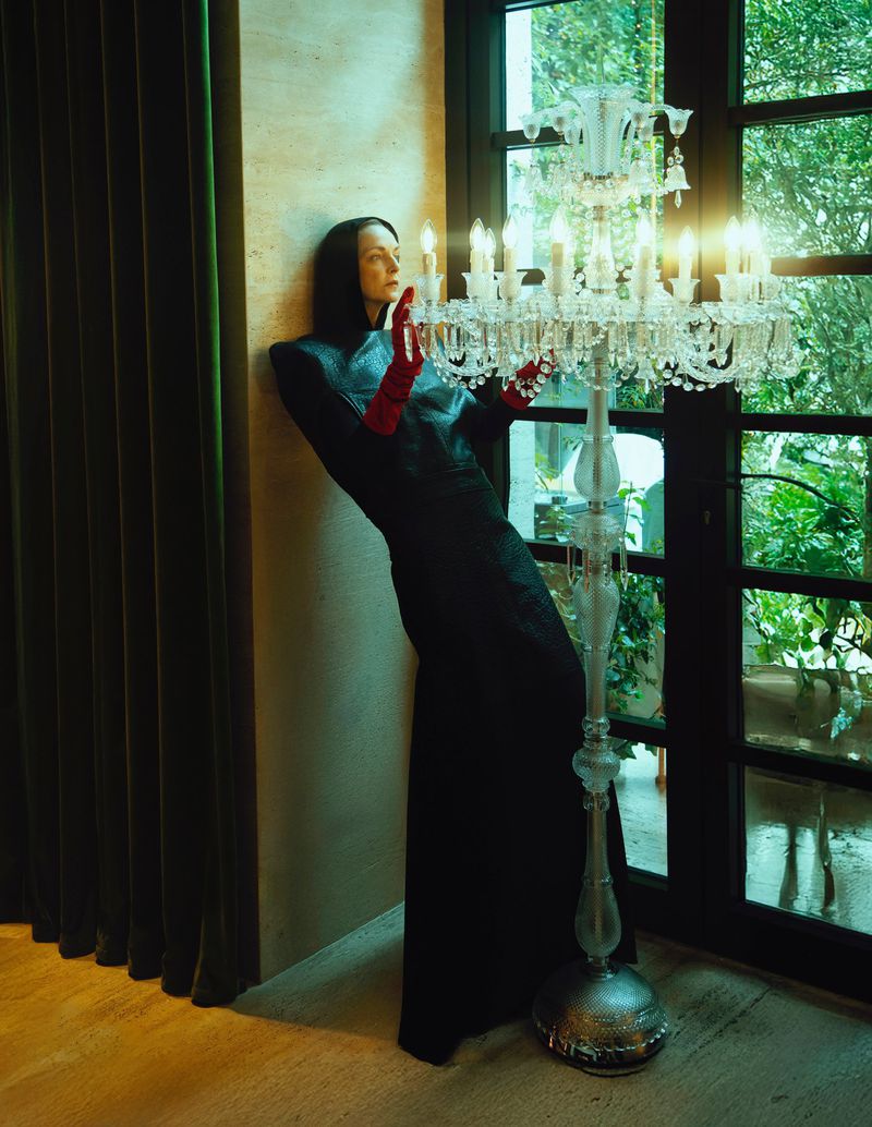 Woman in black dress and red gloves stands next to glass chandelier inside near a window with green foliage visible outside.