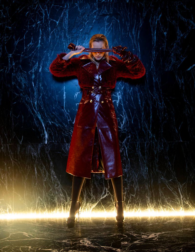 Woman in red vinyl coat poses with a metal rod in a studio with a marble floor and a textured blue-black wall illuminated by yellow light.