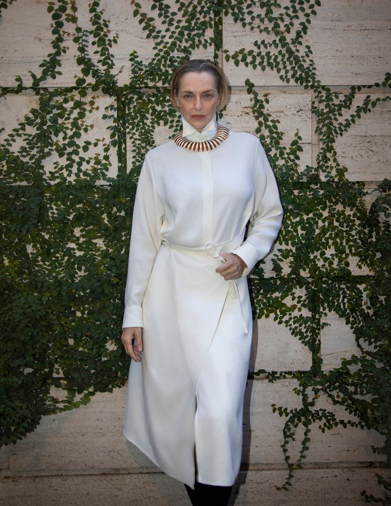 Woman in a white dress and statement necklace posing against a vine-covered wall, showcasing fashion and style outdoors.