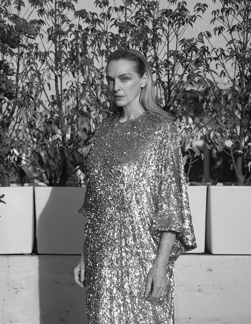 Elegant adult woman in a glittering sequined dress posing outdoors against a leafy green hedge in a black and white portrait.
