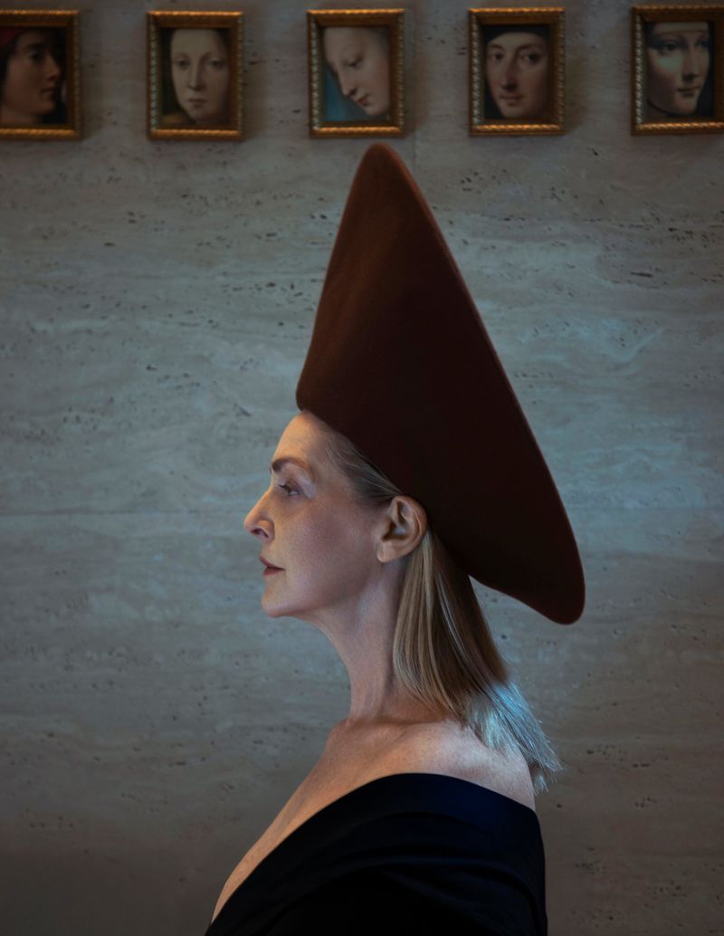 Profile portrait of a mature woman wearing a tall brown hat, with framed portraits hanging on the wall in the background.
