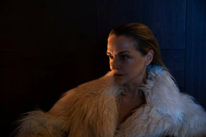 Close-up portrait of an older woman wearing a plush white faux fur collar against a dark wooden wall.
