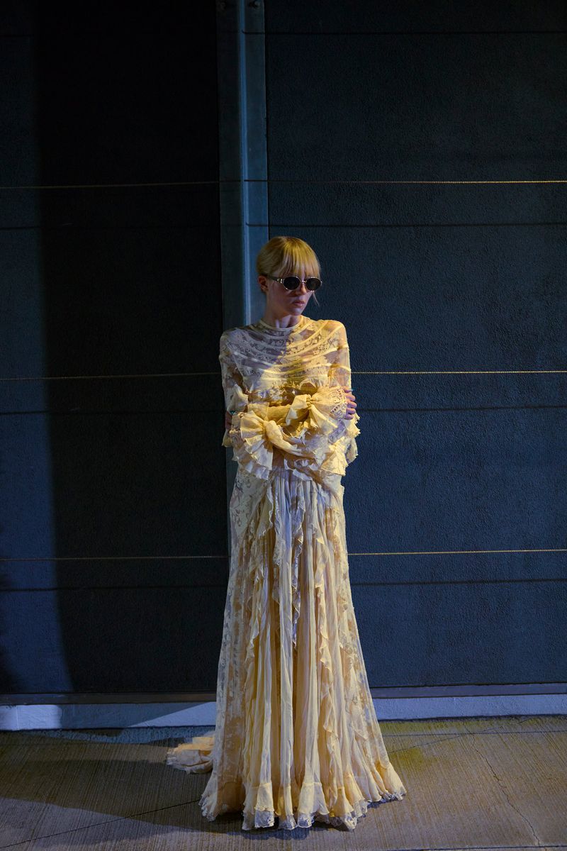 A fair-skinned adult female stands in a long, light yellow ruffled dress and sunglasses against a dark industrial background.