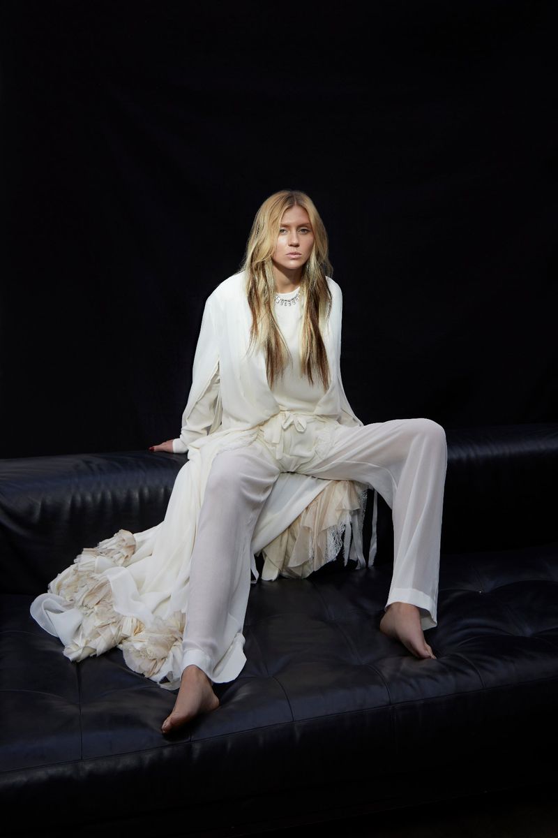 A woman with long blonde hair sits on a black couch wearing a flowing white robe and pants, against a dark backdrop.
