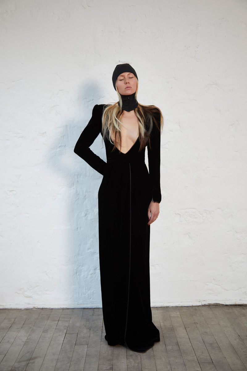 Elegant woman with closed eyes poses in long-sleeved black dress and matching beanie hat against a textured off-white wall in a studio.
