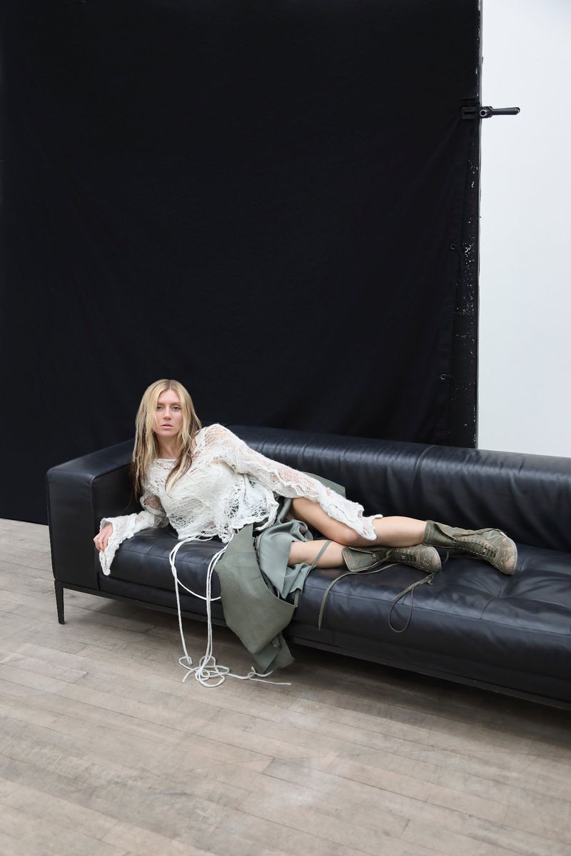 Blond-haired woman lies on a black leather sofa indoors, wearing a lace top and skirt with combat boots in a studio setting.