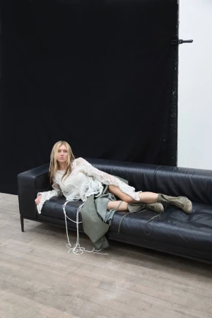 Blond-haired woman lies on a black leather sofa indoors, wearing a lace top and skirt with combat boots in a studio setting.