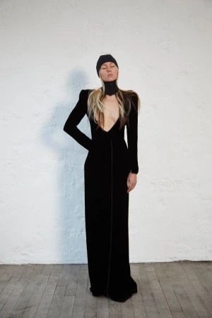 Model wearing a long black dress and black beanie standing against a white textured wall.
