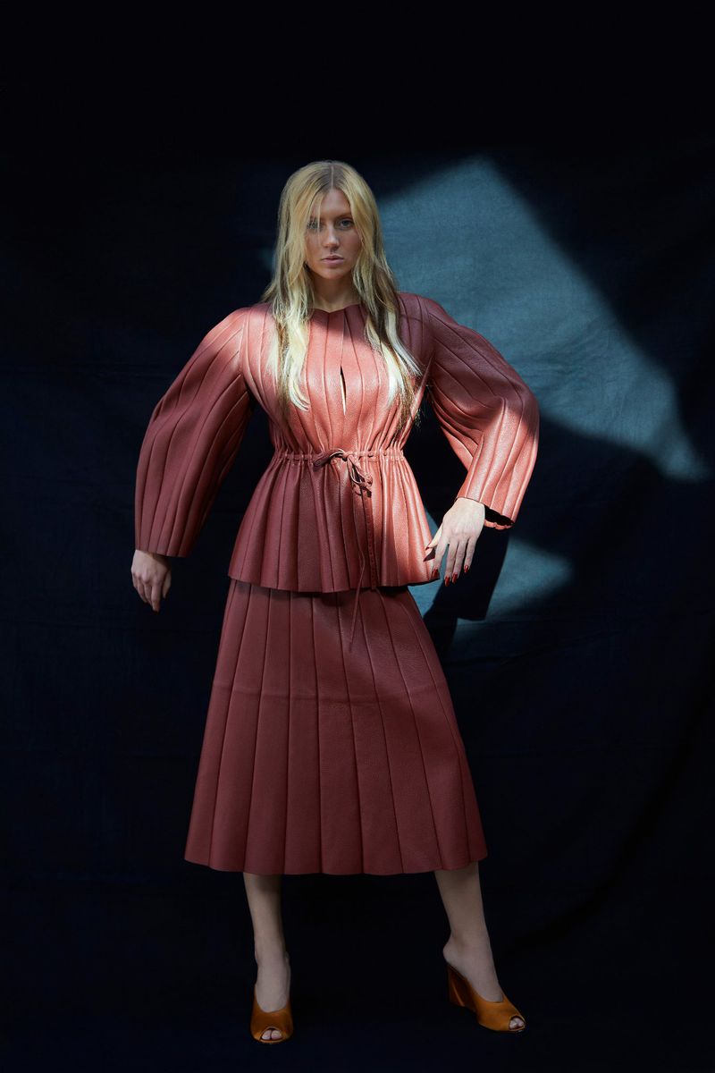 Model wearing a pleated mauve outfit standing against a dark background with dramatic light across her figure.