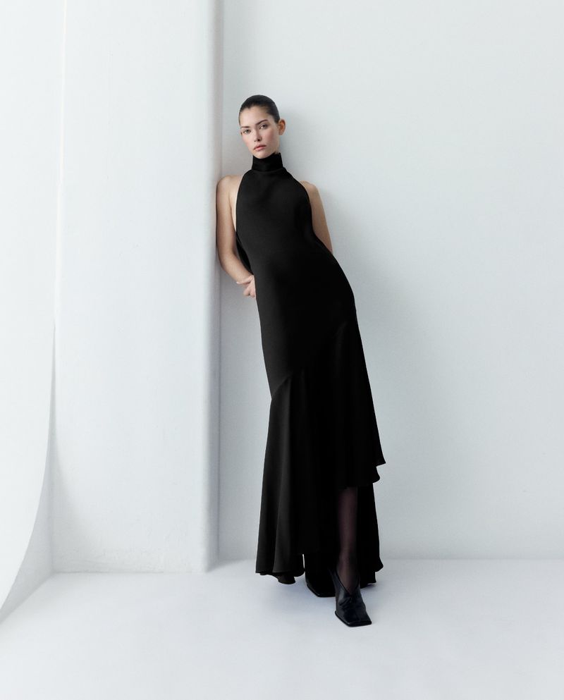 Elegant woman in a black, high-neck gown poses gracefully against a stark white wall, creating a striking contrast in a studio setting.