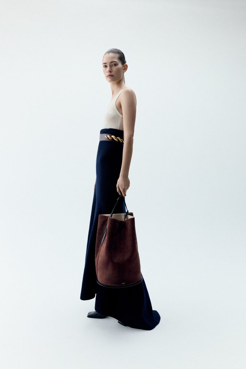 Fashion shot of a woman in a cream-colored top and long blue skirt holding a brown leather bucket bag against a bright white background.