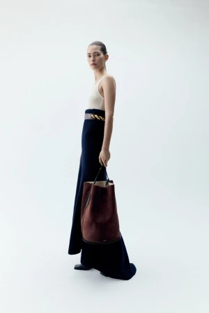 Fashion shot of a woman in a cream-colored top and long blue skirt holding a brown leather bucket bag against a bright white background.