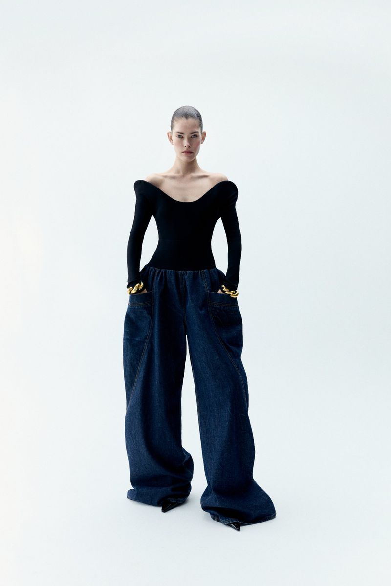 Fashion model wearing a black long-sleeved top and baggy dark blue jeans, posing in a full shot against a white backdrop in a photo studio.