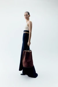 Model wearing a long dark skirt and light top holding a brown suede bucket bag against a white seamless background