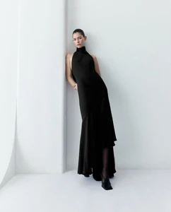 Model in a long black halter-neck dress leaning against a plain white studio wall