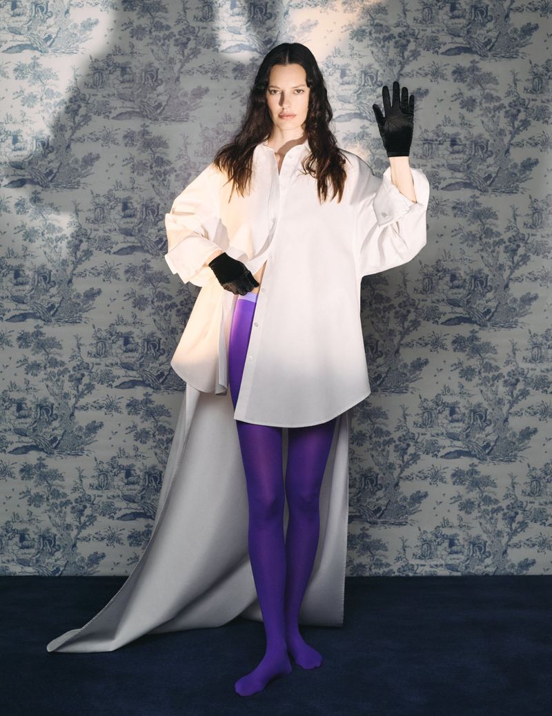Adult woman in a white shirt, purple tights, and black gloves poses in front of a toile-patterned wallpaper with studio lighting.