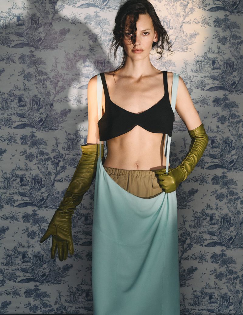 Fashion shot of a young adult woman in bra top, skirt, and long gloves, posed in front of a blue and white patterned wallpaper.